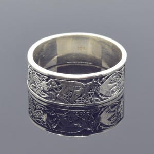 Puede incluir: Un anillo de plata con un diseño intrincado de nudos celtas y criaturas míticas. El anillo es ancho y tiene un acabado pulido.