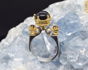 May include: A silver ring with a black gemstone set in a gold crown. The ring has two smaller gold accents with red gemstones on either side of the crown.