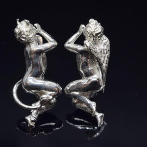 May include: Two silver figurines of angels, one with wings and one with a tail, both with their hands covering their ears.