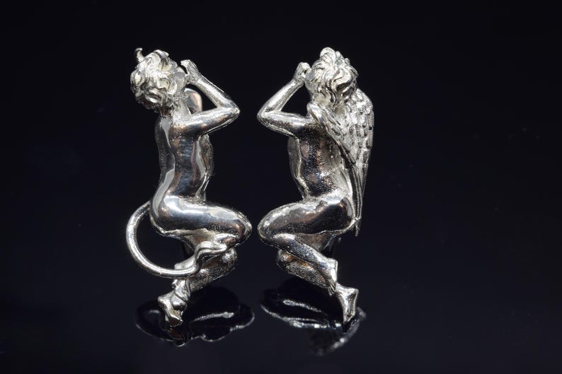 May include: Two silver figurines of angels, one with wings and one with a tail, both with their hands covering their ears.