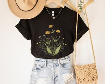 May include: Black t-shirt with a floral print of yellow flowers and green leaves. The shirt is hanging on a wooden hanger with a straw hat and a woven bag.