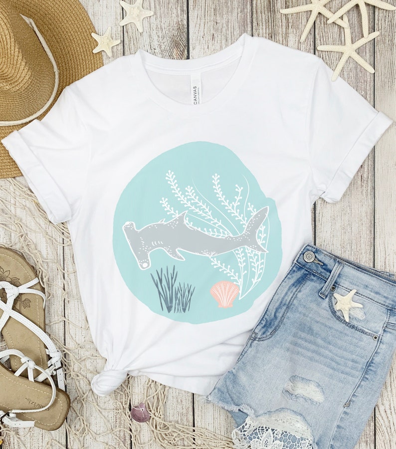 May include: White t-shirt featuring a graphic of a hammerhead shark swimming in a blue circle with seaweed and a seashell. The shirt is laid out on a wooden surface with a straw hat, sandals, and denim shorts.