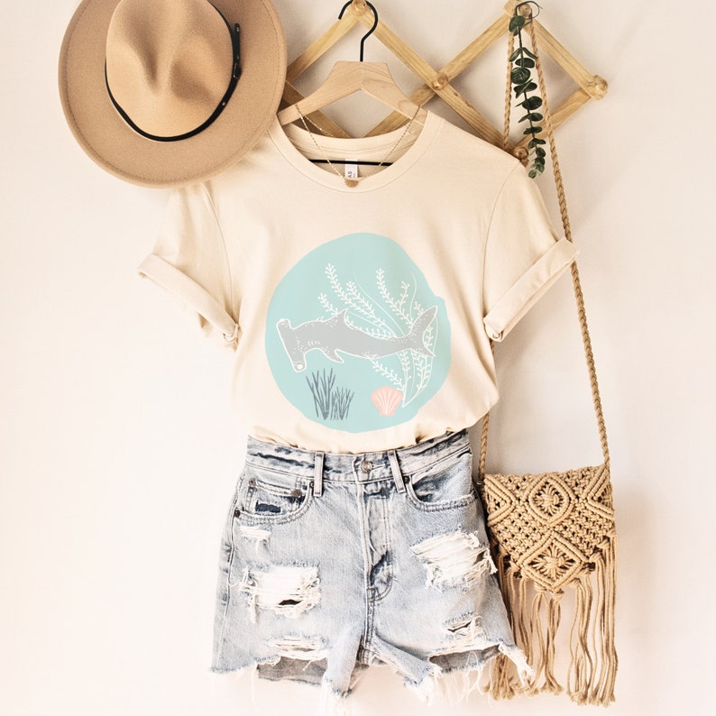 May include: A cream-coloured t-shirt featuring a graphic of a hammerhead shark swimming in a blue circle with seaweed and a seashell. The shirt is paired with light blue denim shorts and a macrame bag.