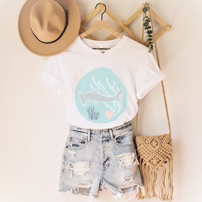 May include: White t-shirt featuring a graphic of a hammerhead shark swimming in a blue circle with seaweed and a seashell. The shirt is hanging on a wooden hanger with a straw hat and a woven bag.