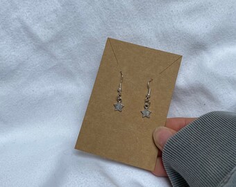 May include: A pair of silver star earrings displayed on a brown card. The earrings are simple and delicate, with a small star charm hanging from a silver hook.