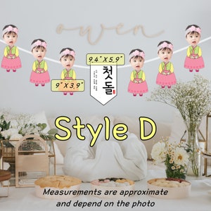 May include: A white banner with a Korean phrase "첫돌" in a red box, and six images of babies in traditional Korean clothing. The banner is decorated with pink flowers and the text "9.4" x 5.9" and "9" x 3.9". The banner is labeled "Style D".