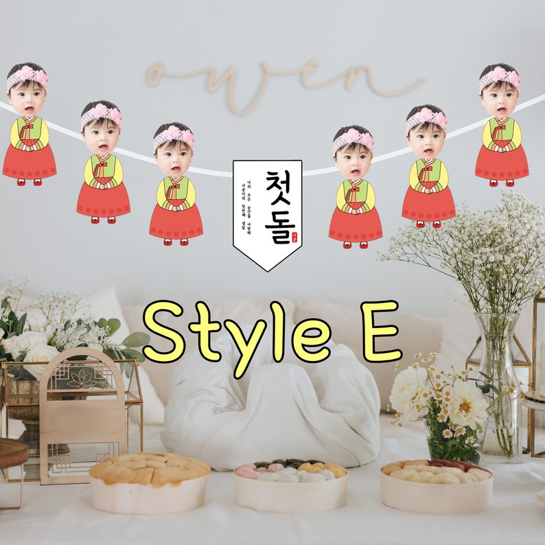 May include: A white banner with a Korean phrase "첫돌" in red and black lettering. The banner is decorated with six images of children wearing traditional Korean clothing. Each child's face is replaced with a photo of a different person. The banner is hanging from a white string.
