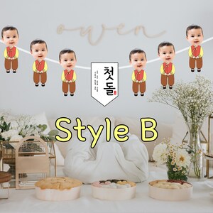 May include: A white banner with a Korean phrase "첫돌" in red and black lettering. The banner is decorated with images of children wearing traditional Korean clothing. The children's faces are replaced with photos. The banner is hanging above a table with a white tablecloth and three bowls of food.