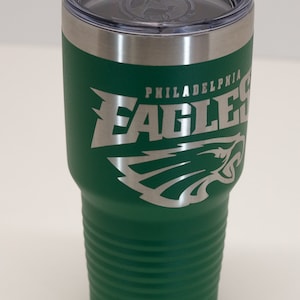 Pu&ograve; includere: Un bicchiere termico verde bosco con il logo e la scritta Philadelphia Eagles incisi in argento sul lato. Il bicchiere presenta una base affusolata con scanalature orizzontali, un bordo in acciaio inossidabile e un coperchio in plastica trasparente.