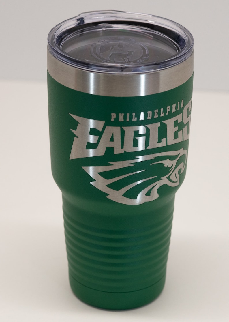 Pu&ograve; includere: Un bicchiere termico verde bosco con il logo e la scritta Philadelphia Eagles incisi in argento sul lato. Il bicchiere presenta una base affusolata con scanalature orizzontali, un bordo in acciaio inossidabile e un coperchio in plastica trasparente.