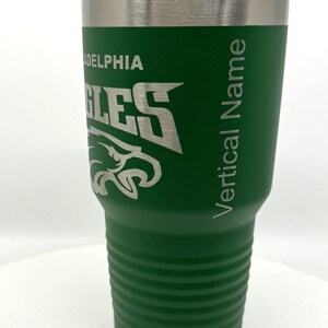 Pu&ograve; includere: Un bicchiere in acciaio inossidabile verde con coperchio trasparente, caratterizzato da un logo inciso dei Philadelphia Eagles e dal testo Vertical Name sul lato. Il bicchiere ha un design affusolato con scanalature orizzontali sulla base.