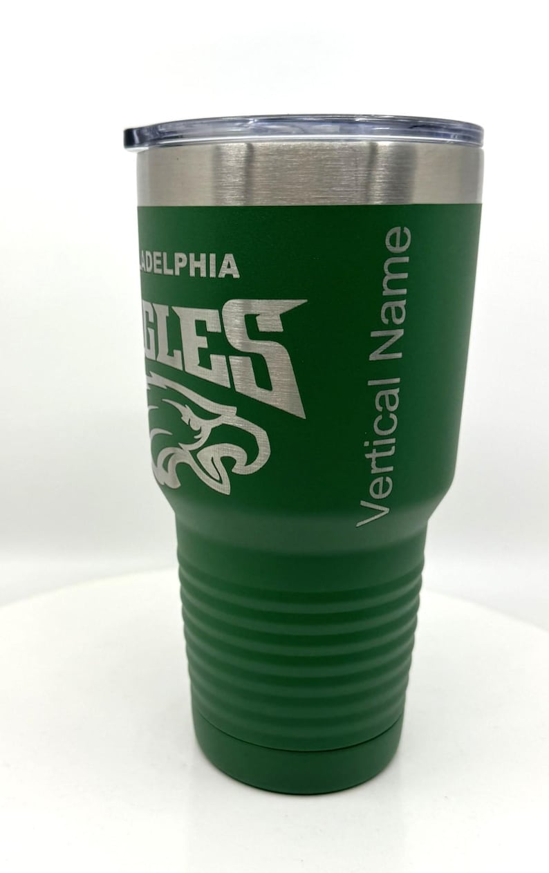 Pu&ograve; includere: Un bicchiere in acciaio inossidabile verde con coperchio trasparente, caratterizzato da un logo inciso dei Philadelphia Eagles e dal testo Vertical Name sul lato. Il bicchiere ha un design affusolato con scanalature orizzontali sulla base.