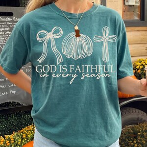 Peut inclure: Un t-shirt bleu sarcelle à manches courtes avec une impression graphique blanche représentant un nœud, une citrouille et une croix. Le texte "GOD IS FAITHFUL in every season" est imprimé en blanc au centre. Le vêtement a un col rond.