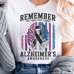Może przedstawiać: Biały t-shirt z grafiką zawierającą napis REMEMBER FOR THOSE THAT CANNOT ALZHEIMER'S AWARENESS. Wzór przedstawia mózg podzielony na różową i niebieską połowę, czarną wstążkę świadomości oraz tło z wyblakłą flagą Stanów Zjednoczonych.