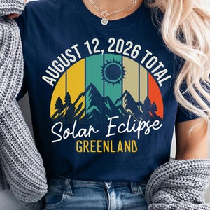 Könnte beinhalten: Ein marineblaues T-Shirt mit einer Grafik einer Sonnenfinsternis über Bergen und dem Text AUGUST 12, 2026 TOTAL Solar Eclipse GREENLAND. Das Design zeigt eine retro-artige Sonne und eine Bergsilhouette in gelben, grünen und orangen Streifen.