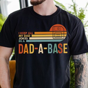 May include: Black t-shirt with the text "I KEEP ALL MY DAD JOKES IN A DAD-A-BASE" in a retro design. The design includes a floppy disk graphic and a sunset-style color scheme. A casual, comfortable tee.