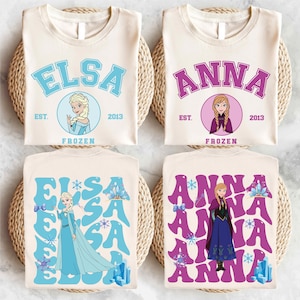 Könnte beinhalten: Vier weiße T-Shirts mit verschiedenen Designs, die die Figuren Elsa und Anna aus dem Film Frozen zeigen. Die Shirts haben den Text "Elsa" und "Anna" in einer College-Schriftart mit dem Text "Frozen" und dem Jahr "2013" darunter. Zwei der Shirts haben ein einfacheres Design mit einem Bild von Elsa und Anna in einem Kreis. Die anderen beiden Shirts haben ein komplexeres Design mit den Namen "Elsa" und "Anna", die mehrmals in einer Retro-Schriftart wiederholt werden.