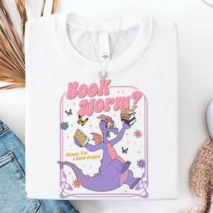 a white t-shirt with a colorful graphic design depicting a purple dragon holding a book and surrounded by butterflies and flowers.