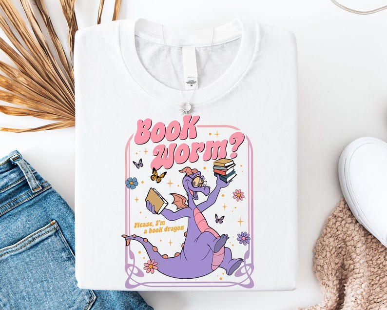 a white t-shirt with a colorful graphic design depicting a purple dragon holding a book and surrounded by butterflies and flowers.