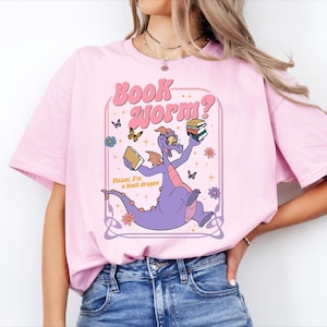 A woman wearing a pink t-shirt with a graphic of a purple dragon holding a book and surrounded by butterflies and flowers.