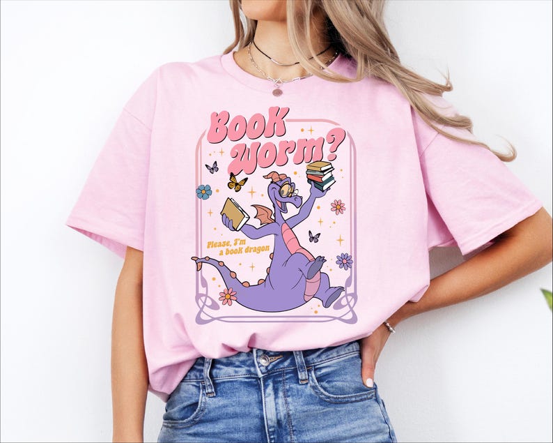 A woman wearing a pink t-shirt with a graphic of a purple dragon holding a book and surrounded by butterflies and flowers.