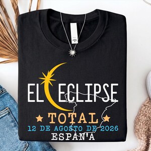 Op de afbeelding: Een zwart t-shirt met een grafisch ontwerp van een gele maansikkel en ster, een witte omtrek van Spanje en de tekst EL ECLIPSE TOTAL 12 DE AGOSTO DE 2026 ESPAÑA. Het t-shirt heeft een ronde hals en een wit label in de kraag.