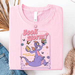 A pink t-shirt with a cartoon dragon holding a book and surrounded by butterflies and flowers, along with a pair of jeans and a white sweater.