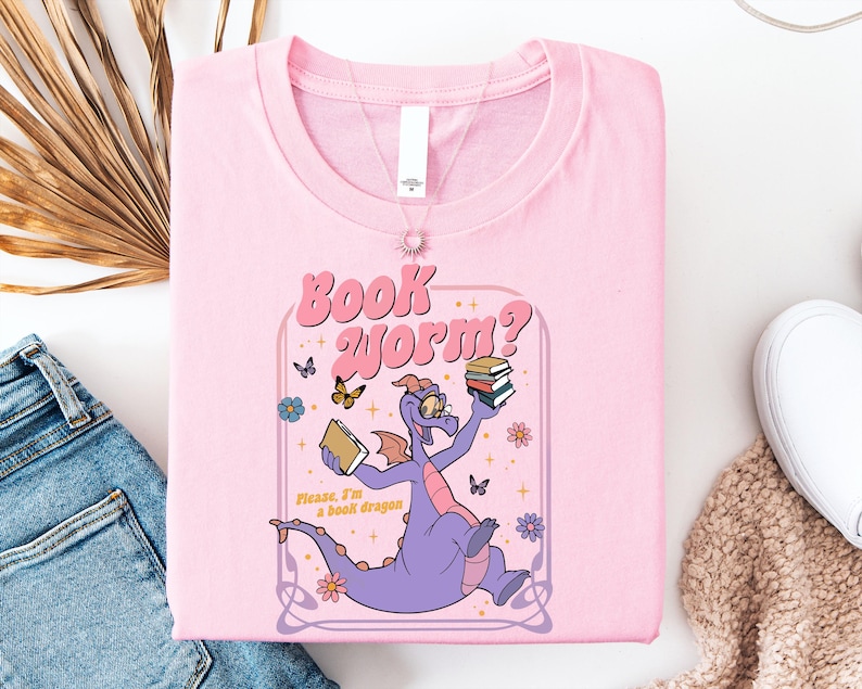 A pink t-shirt with a cartoon dragon holding a book and surrounded by butterflies and flowers, along with a pair of jeans and a white sweater.