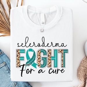 Könnte beinhalten: Ein weißes T-Shirt mit dem schwarzen Schriftzug "scleroderma" über dem Wort "FIGHT" in großen Blockbuchstaben mit einem blaugrünen Leopardenmuster sowie dem Text "For a cure" darunter, ergänzt durch eine integrierte blaugrüne Schleife.