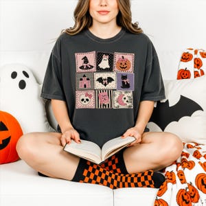 Op de afbeelding: Een antracietgrijs T-shirt met korte mouwen voorzien van een raster van negen vierkante vlakken met Halloween-illustraties, waaronder een heksenhoed, spook, pompoen, spookhuis, vleermuis, zwarte kat, schedel, spreukenboek en een halve maan.