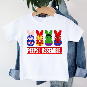 May include: White t-shirt featuring four cartoon bunny characters dressed as superheroes. The bunnies are in red, blue, green, and yellow. The text "PEEPS! ASSEMBLE." is printed in red on a white rectangle.