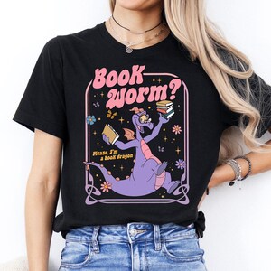 A woman wearing a black t-shirt with a graphic design featuring a purple dragon holding books and surrounded by stars and flowers.