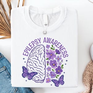 Może przedstawiać: Biały t-shirt z grafiką i napisem EPILEPSY AWARENESS. Ilustracja przedstawia mózg podzielony na dwie połowy, z których jedna ma teksturę mózgu, a druga składa się z fioletowych kwiatów, całość uzupełniają dwa fioletowe motyle.