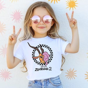 A young girl wearing a white t-shirt with a graphic of a cartoon character and pink sunglasses is making a peace sign with her hands.