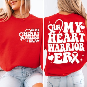 Puede incluir: Sudadera roja con texto blanco que dice "In My Heart Warrior Era" con una cinta blanca y un corazón. La parte posterior de la sudadera tiene el mismo texto en una fuente más grande con una cinta blanca.