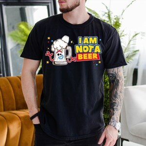 A man wearing a black t-shirt with a graphic design of a beer mug and the text "I AM NOT A BEER" is standing in a room with a couch and a potted plant.