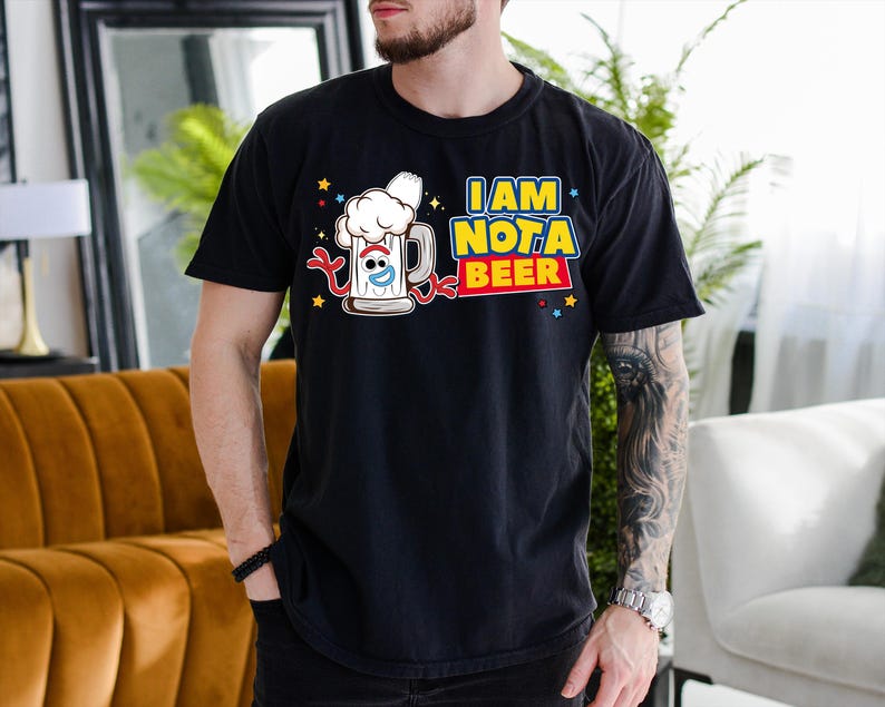 A man wearing a black t-shirt with a graphic design of a beer mug and the text "I AM NOT A BEER" is standing in a room with a couch and a potted plant.