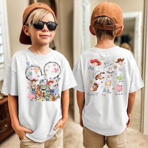 A young boy wearing a white t-shirt with a cartoon character design and sunglasses, standing in front of a mirror.