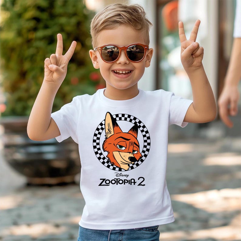 A young boy wearing sunglasses and a white t-shirt with a cartoon character graphic is smiling and making a peace sign gesture.