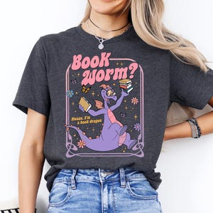 A woman wearing a gray t-shirt with a graphic design of a purple dragon holding a book and surrounded by flowers and butterflies.