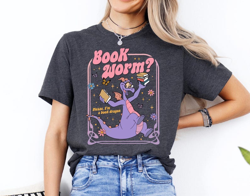 A woman wearing a gray t-shirt with a graphic design of a purple dragon holding a book and surrounded by flowers and butterflies.