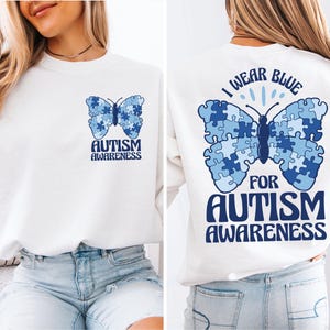 Puede incluir: Sudadera blanca con una mariposa azul hecha de piezas de rompecabezas. La mariposa tiene el texto "Uso azul por la concienciación sobre el autismo" en la espalda. La parte delantera de la sudadera tiene una mariposa azul más pequeña con el texto "Concienciación sobre el autismo".