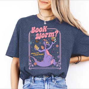 A woman wearing a blue t-shirt with a graphic design featuring a purple dragon holding a book and surrounded by butterflies and flowers.