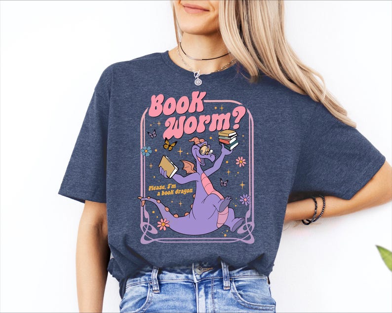A woman wearing a blue t-shirt with a graphic design featuring a purple dragon holding a book and surrounded by butterflies and flowers.