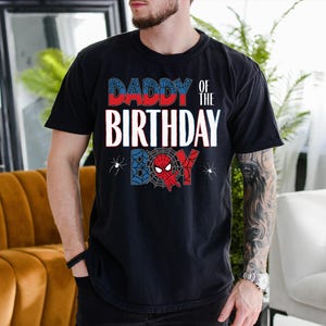 A man wearing a black t-shirt with a Spider-Man design and text that says "DADBY OF THE BIRTHDAY" is standing in a room with a couch and a plant.