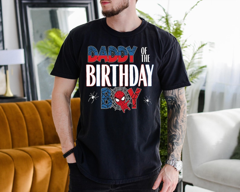 A man wearing a black t-shirt with a Spider-Man design and text that says "DADBY OF THE BIRTHDAY" is standing in a room with a couch and a plant.