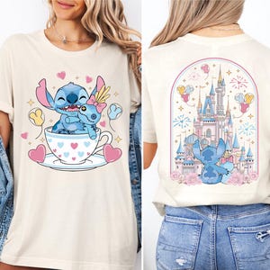 A woman wearing a white t-shirt with a graphic design featuring a blue character, likely Stitch, sitting in a teacup with hearts and a castle in the background.