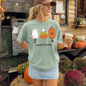 Peut inclure: Un t-shirt à manches courtes vert clair avec un motif représentant trois sucettes glacées sur le thème d'Halloween : un fantôme blanc, un bonbon maïs jaune et orange, et une citrouille orange. Le texte "Sweet & Spooky!" est imprimé en noir.