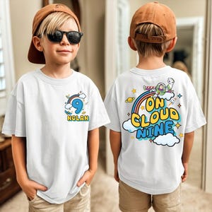 A young boy wearing a white t-shirt with a graphic design and text on the back, standing in front of a mirror.