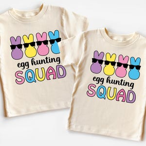 Könnte beinhalten: Zwei beigefarbene T-Shirts mit einem Motiv von vier pastellfarbenen Osterhasen, die Sonnenbrillen tragen. Der Text auf den Shirts lautet "egg hunting SQUAD".